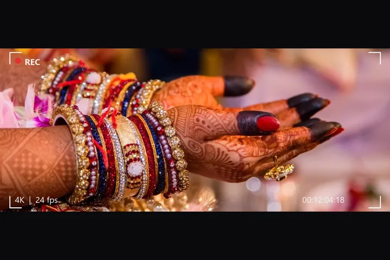 A wedding film frame in a cinematic letterbox with timecode, bride's hands with henna and bangles
